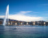 Drohnenaufnahme von Genf vom See aus, die den Jet-d-Eau und die genfer Küste zeigt. Im Vordergrund fährt ein Boot richtung Stadt.