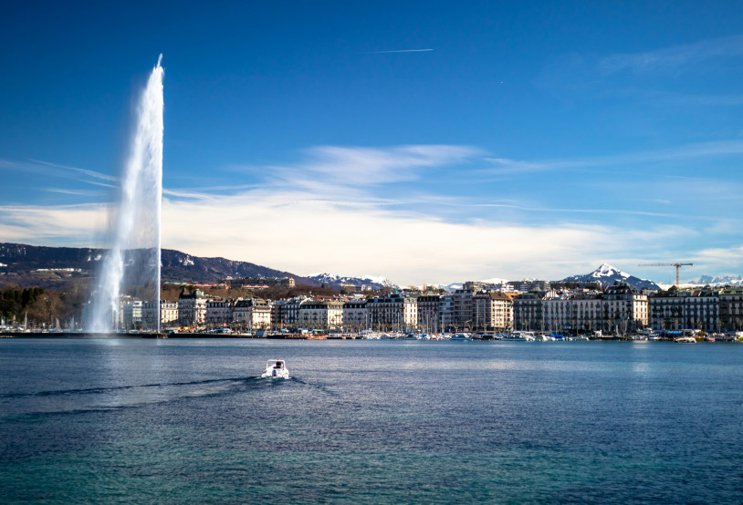 Drohnenaufnahme von Genf vom See aus, die den Jet-d-Eau und die genfer Küste zeigt. Im Vordergrund fährt ein Boot richtung Stadt.