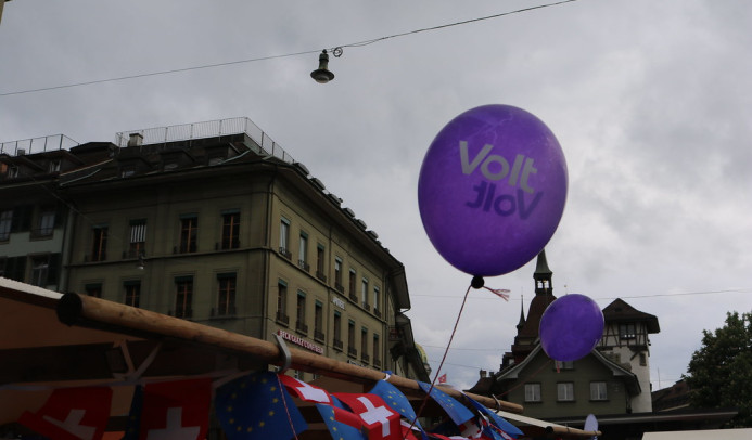 Zwei lilafarbene Luftballons mit weißem „Volt“-Schriftzug schweben an Schnüren über dem Holzdach eines Marktstands vor einem bewölkten Himmel. Am Rand des Standes hängt eine Wimpelkette aus abwechselnd angeordneten Schweizer- und Europaflaggen. Im Hintergrund sind historische Altstadtgebäude mit Spitztürmen sowie über die Straße gespannte Oberleitungen und eine hängende Straßenlaterne zu sehen.