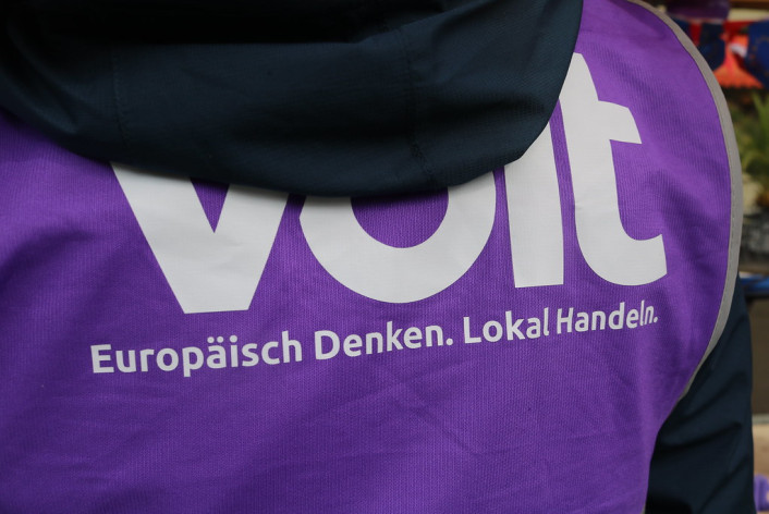 Nahaufnahme des Rückens einer Person, die über einer dunkelblauen Jacke eine leuchtend violette Kampagnenweste trägt. Auf der Weste ist in großen weißen Buchstaben der Schriftzug „Volt“ aufgedruckt, der oben teilweise von der Kapuze verdeckt wird, darunter der weiße Slogan: „Europäisch Denken. Lokal Handeln.“