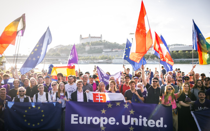 Eine große, gut gelaunte Gruppe von Unterstützerinnen und Unterstützern der pro-europäischen Partei „Volt“ posiert für ein Gruppenfoto am Ufer der Donau in Bratislava, Slowakei, mit der historischen Burg Bratislava im Hintergrund auf einem Hügel. Im Vordergrund halten sie ein großes, dunkelblaues Banner mit gelben EU-Sternen und dem weißen Schriftzug „Europe. United.“, flankiert von einer EU-Flagge und einer slowakischen Flagge. Zahlreiche Menschen im dichten Gedränge lächeln, winken und schwenken eine Vielzahl von Flaggen, darunter EU-Fahnen, Nationalflaggen (wie die von Slowakei, Österreich, Schweiz und Belgien) sowie lilafarbene „Volt“-Fahnen. Viele tragen T-Shirts mit dem „Volt“-Logo oder violette Accessoires. Das Bild ist von warmem, natürlichem Tageslicht beleuchtet.