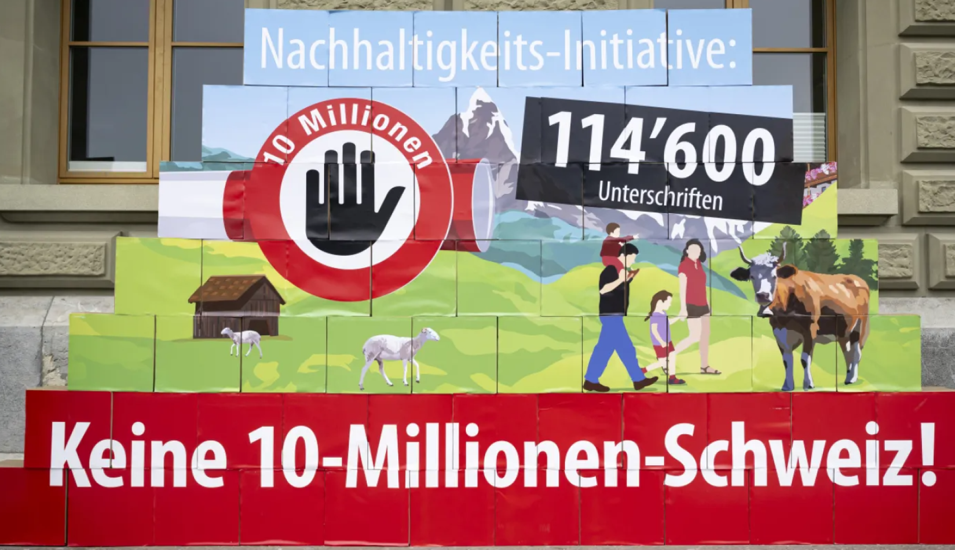 Eine Pyramide aus bedruckten Boxen vor einer Steinfassade zeigt eine ländliche Schweizer Landschaft mit Bergen, einer Familie, einer Kuh und Schafen. Darauf integriert sind ein Stoppsymbol mit einer Hand und dem Text «10 Millionen» sowie die Schriftzüge «Nachhaltigkeits-Initiative:», «114'600 Unterschriften» und gross auf rotem Grund am Fuss der Pyramide «Keine 10-Millionen-Schweiz!».