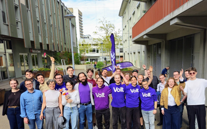 Eine Gruppe von etwa 20 gut gelaunten Menschen posiert im Freien auf einem asphaltierten Platz vor modernen Gebäuden. Mehrere Personen in der Mitte tragen violette T-Shirts oder Hoodies mit dem weissen Aufdruck «Volt». Einige Personen halten Blumen hoch oder haben die Arme jubelnd in die Luft gestreckt. Im Hintergrund ist eine violette Volt-Fahne zu sehen. Das Bild vermittelt eine positive, gemeinschaftliche Atmosphäre.