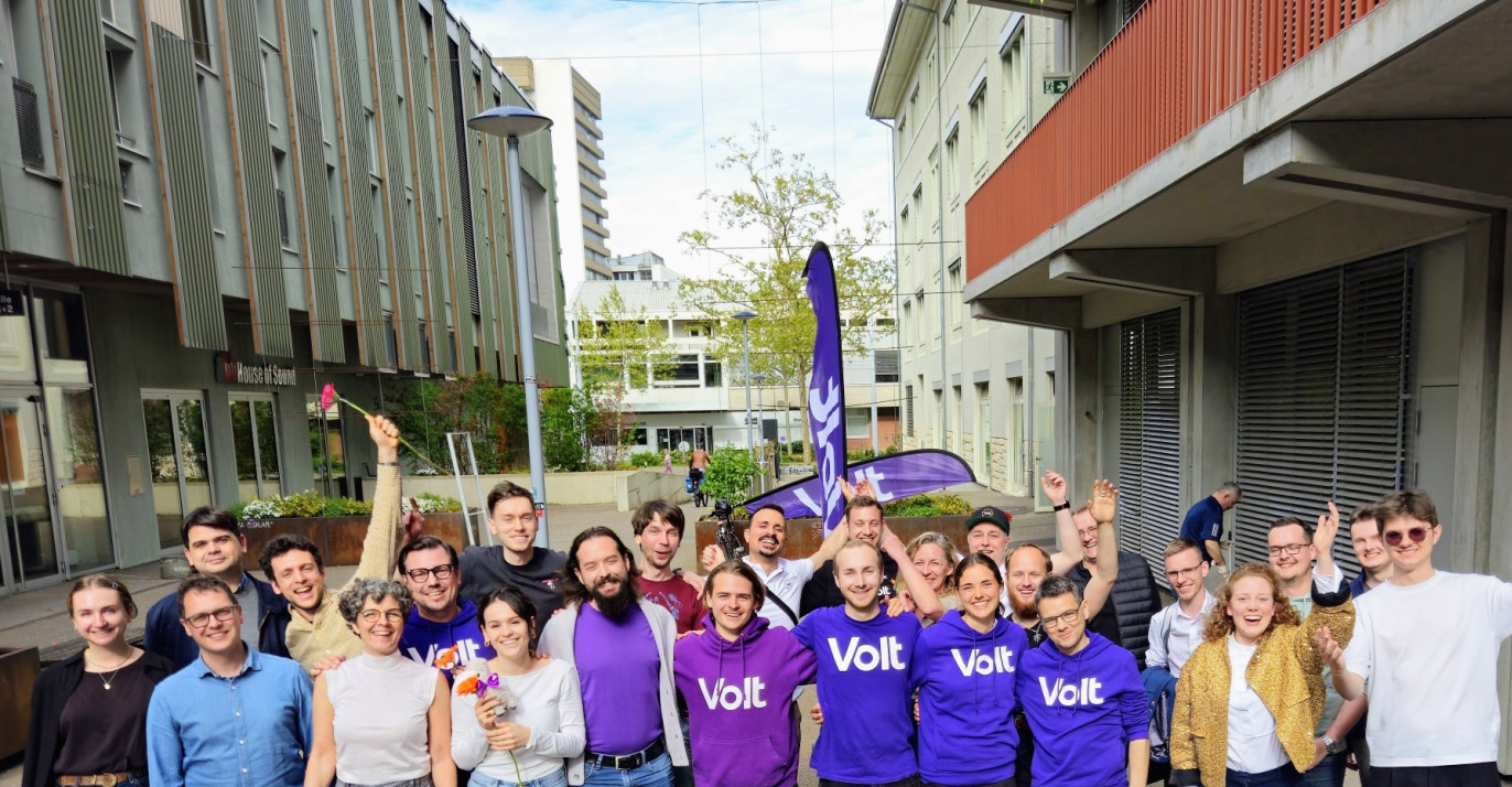 Eine Gruppe von etwa 20 gut gelaunten Menschen posiert im Freien auf einem asphaltierten Platz vor modernen Gebäuden. Mehrere Personen in der Mitte tragen violette T-Shirts oder Hoodies mit dem weissen Aufdruck «Volt». Einige Personen halten Blumen hoch oder haben die Arme jubelnd in die Luft gestreckt. Im Hintergrund ist eine violette Volt-Fahne zu sehen. Das Bild vermittelt eine positive, gemeinschaftliche Atmosphäre.